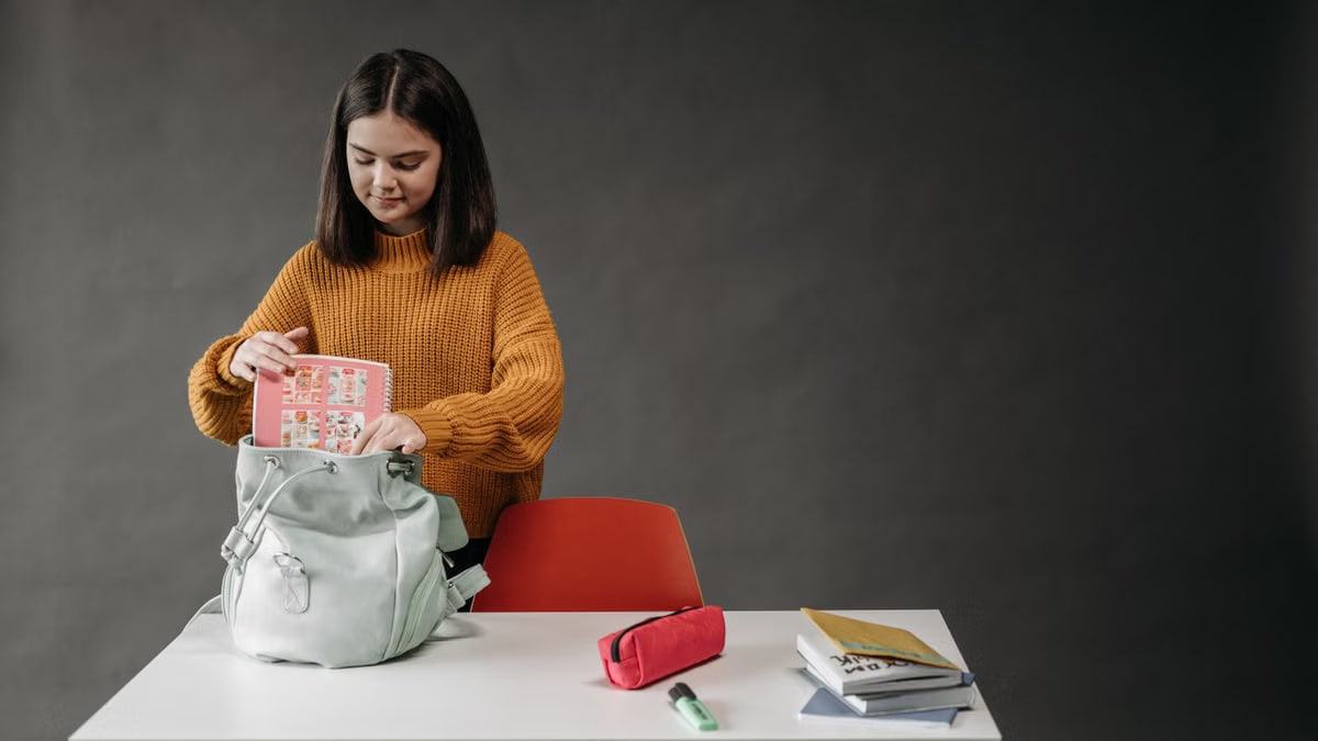 Caucasian girl organizing school supplies into her backpack at a desk.